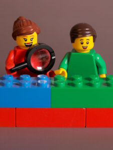 Two smiling LEGO minifigures stand behind a colorful stack of LEGO bricks in blue, green, and red. The woman minifigure, wearing a red shirt, holds a magnifying glass, while the man minifigure next to her is dressed in a green shirt.