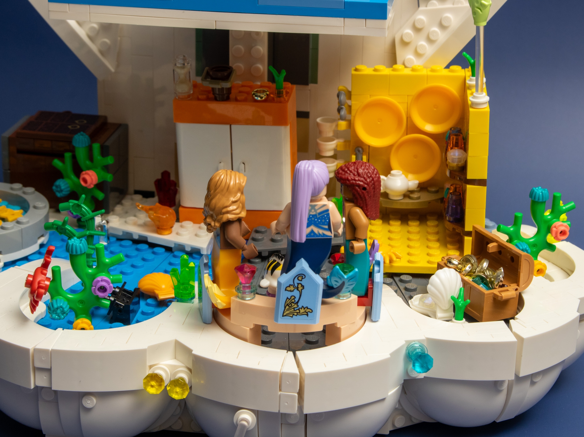 A close-up view of a modified LEGO Royal Clamshell set transformed into a Polly Pocket-style house. In the foreground, three mermaid minifigures are seated on a couch. Behind them is a wardrobe and a kitchen. Additional elements include a treasure chest and plants. 